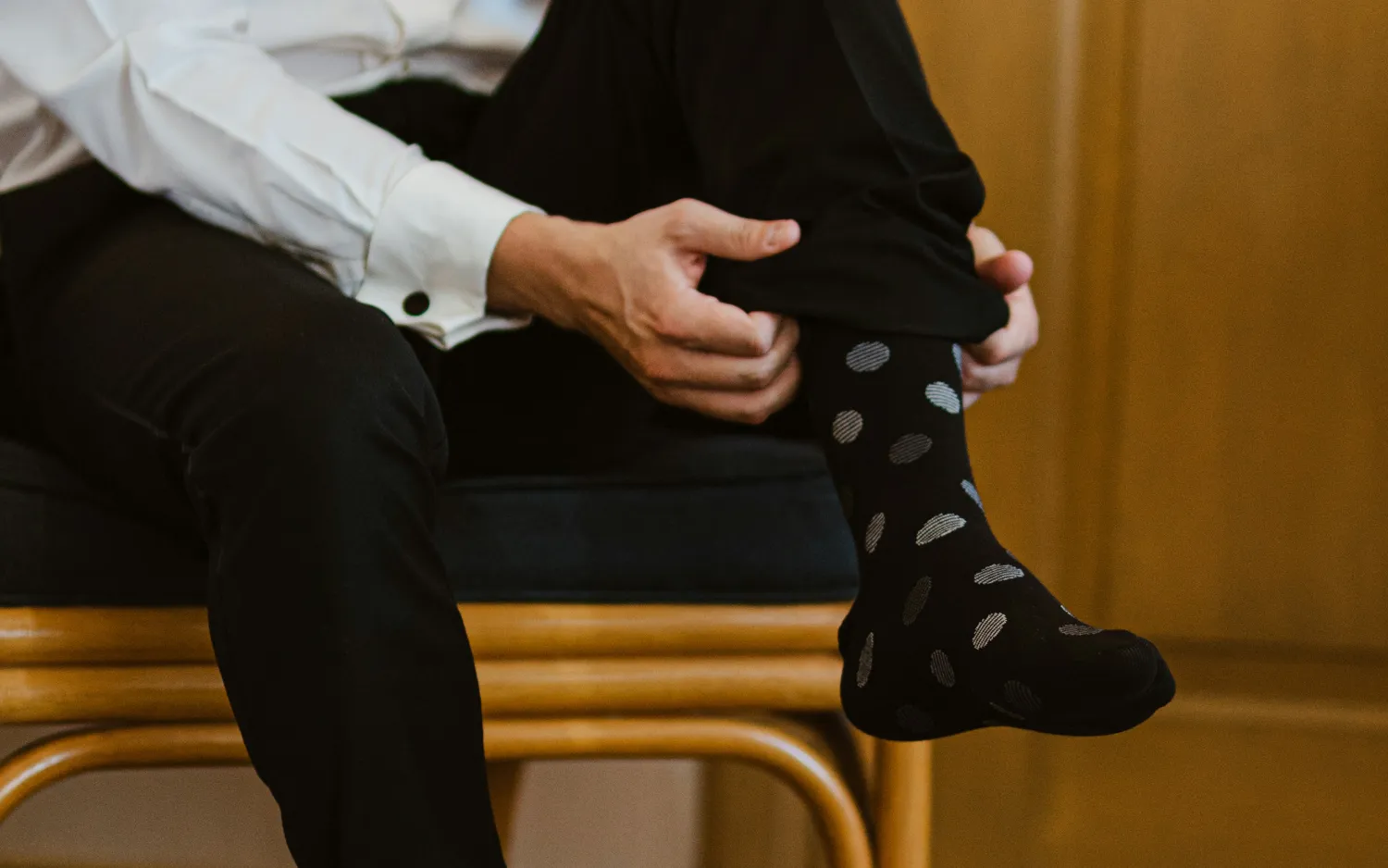 Man in white dress shirt and cufflinks pulling on a black polka-dot sock, dress shoes nearby