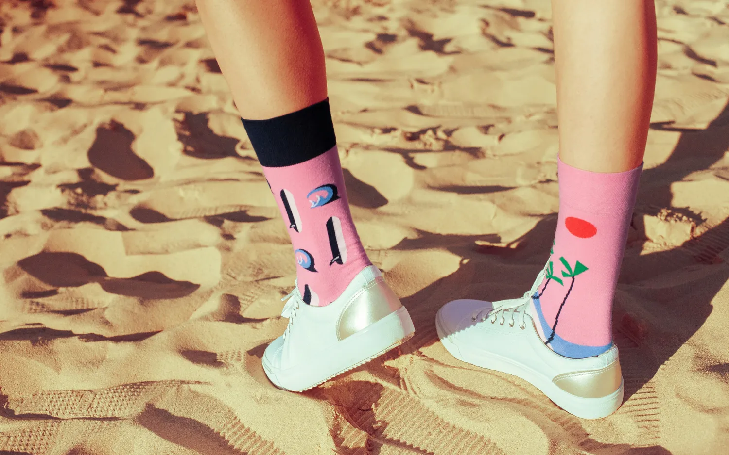 Pink designer socks with abstract art prints worn with white canvas sneakers on a sandy beach
