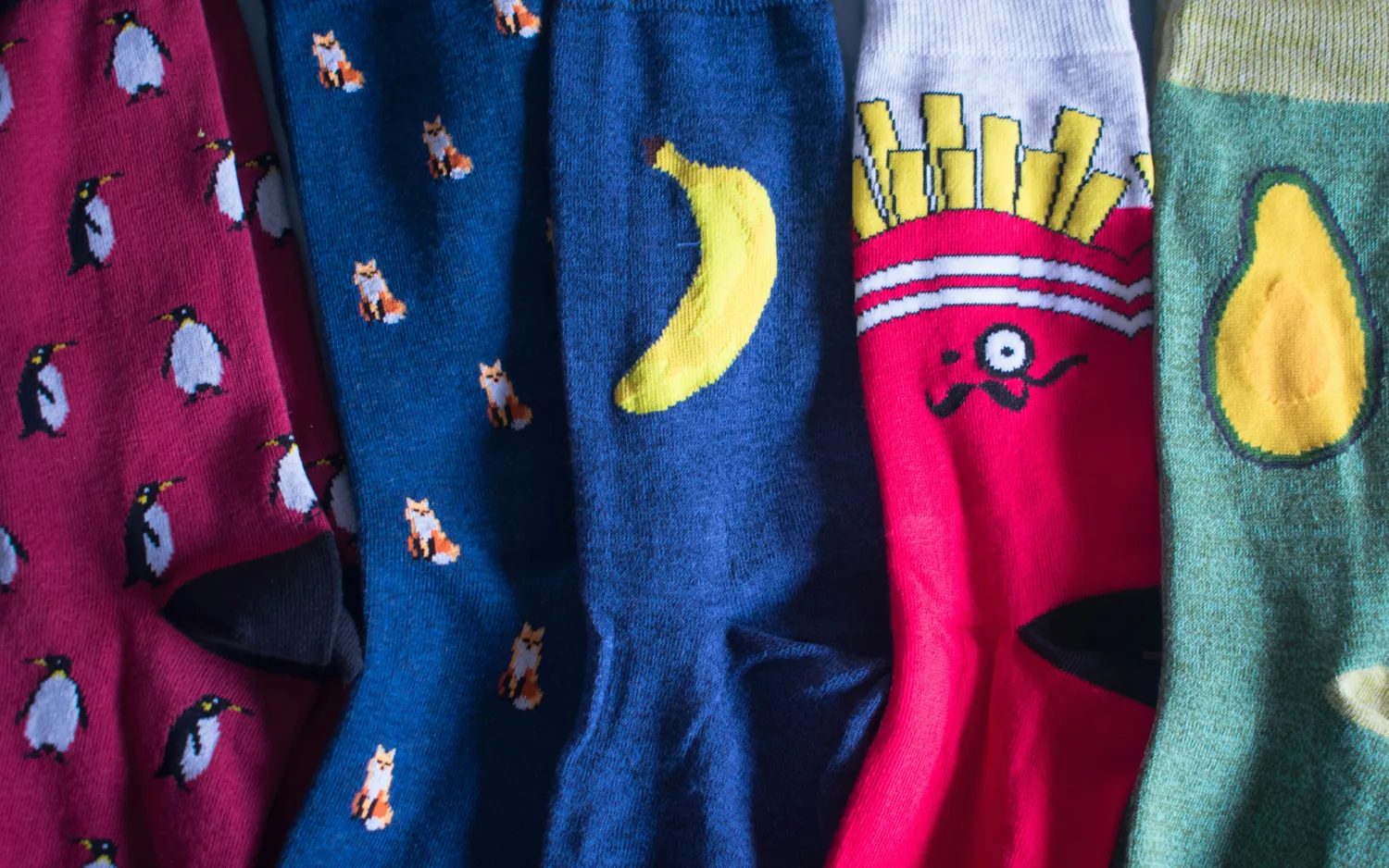Five custom-knit socks side by side with intricate character motifs: penguins, foxes, banana, fries character, and avocado